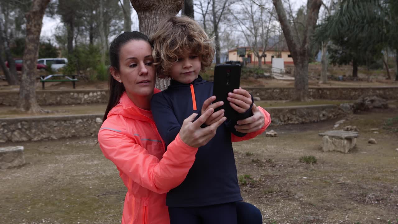 mujer deportiva sonriente con su hijo tomando una selfie en el teléfono inteligente al aire libre