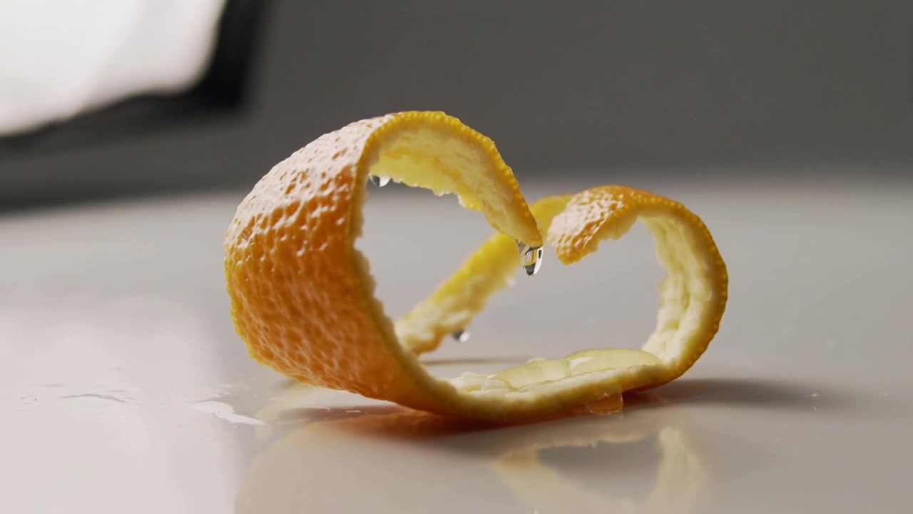 Eine tropfende Herz-Orangenschale gibt Tropfen ab, die als wachsende und fallende Perlen auf einer glänzenden weißen Arbeitsplatte erscheinen.