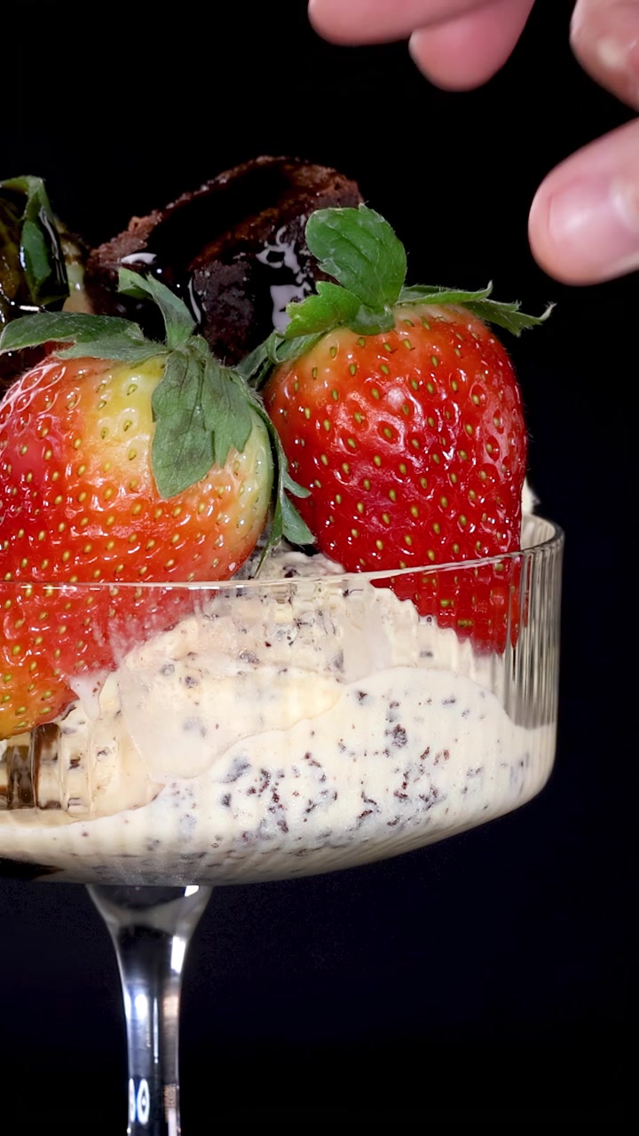 Une main ajoute délicatement une fraise mûre à un dessert en verre composé de glace à la vanille, de sauce au chocolat, de morceaux de brownie et de pépites de chocolat, sous un éclairage de studio.