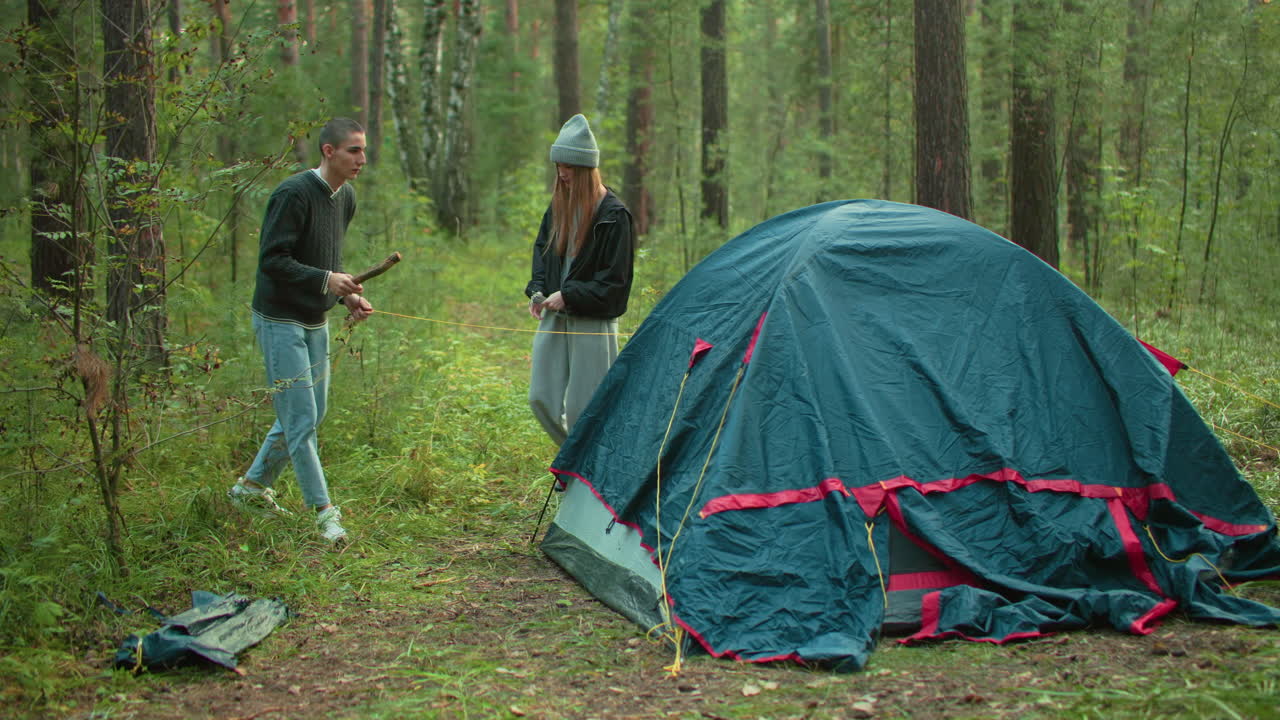 Man grabs yellow tent rope preparing to peg it into ground as woman stands nearby watching in peaceful forest, both actively involved in setting up tent during camping trip surrounded by nature