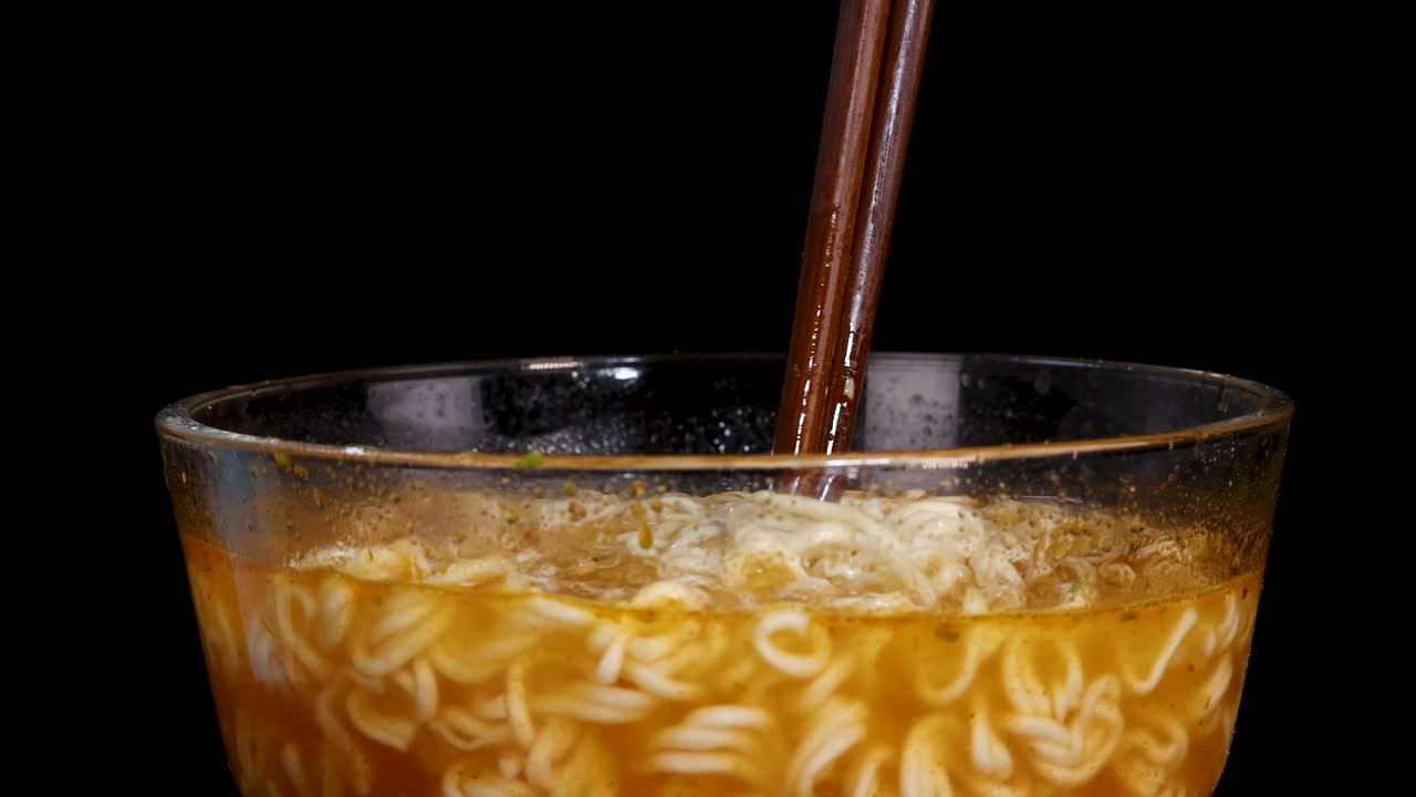 A pair of chopsticks stirs steaming instant noodles in a clear glass bowl against a black background, with even studio lighting and minimal camera movement