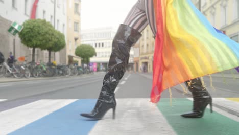 primer plano de las piernas de una transgénero con largos tacones negros cruzando un paseo cruzado lgbtq arcoíris en un entorno urbano, usando una bufanda arcoíris