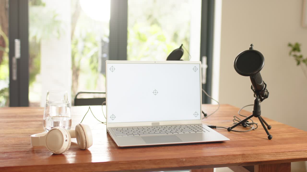 Camera starting capturing desk with laptop, headphones, water glass, microphone in daylight