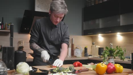 Young-professional-male-chef-in-an-elegant-black-shirt-with-an-alternative-look,-tattoos,-gloves-chopping-red-peppers-A-modern,-fancy-looking-kitchen-with-fresh-vegetables-besides-Cinematic-Sliding-4K