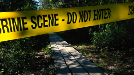 yellow vibrant colored police crime scene do not enter tape pulled over and in front of a wooden boardwalk in a forest marshland bog or wetlands during a sunny day with trees and bushes on the sides