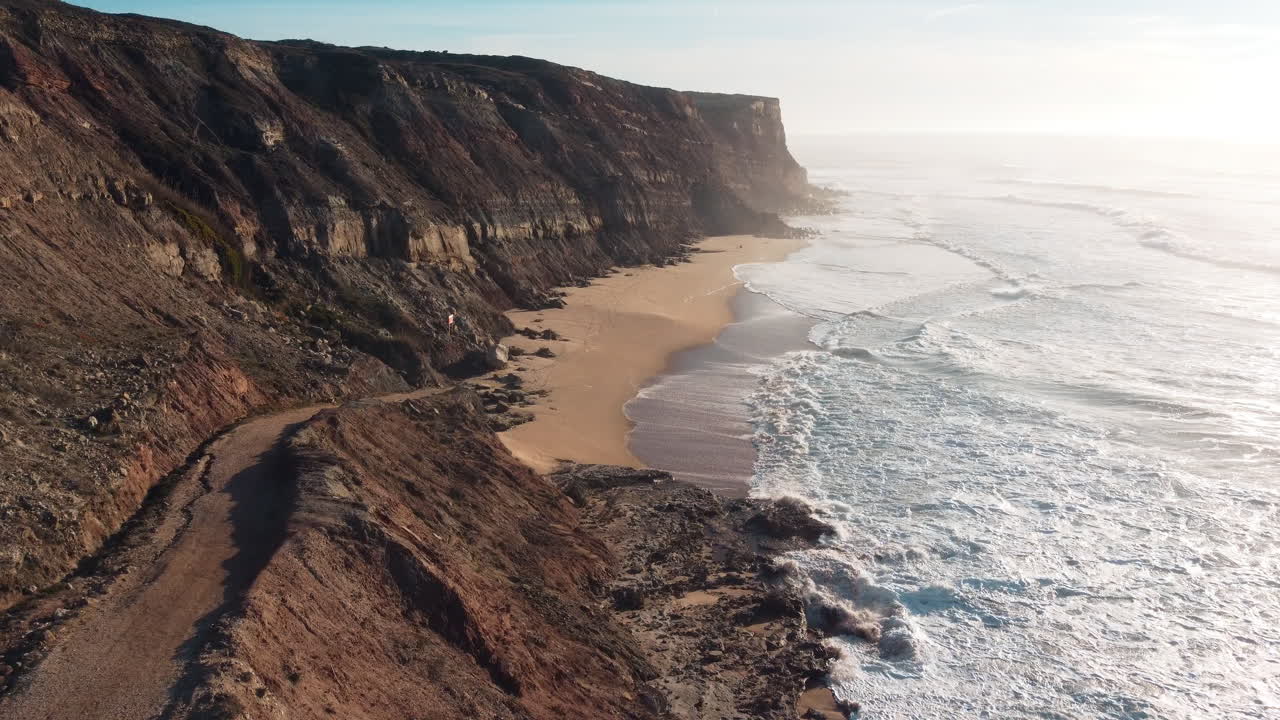 Cinematic drone flight along coastline of Portugal with rocky cliffs and foaming waves of ocean at sunset