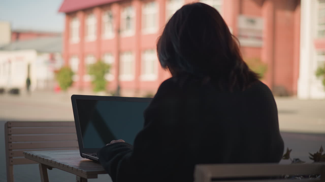 Woman working on laptop outdoors with hair partially covering face, laptop screen reflecting light, urban setting features blurred red brick building and greenery