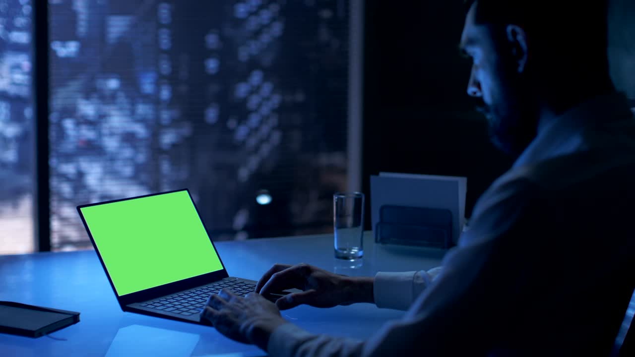 Late at Night Businessman Works on a Laptop with Green Mock-up Screen in His Private Office with Big City Window View.
