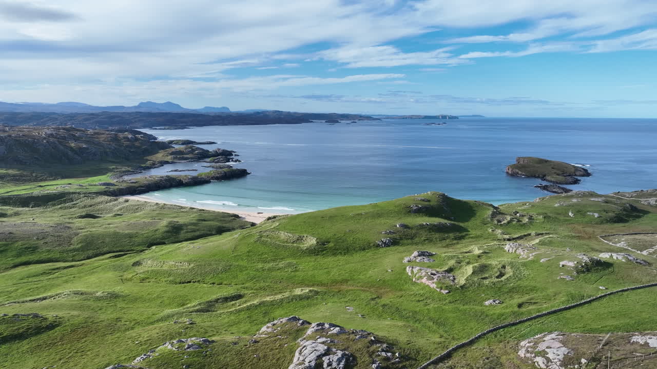 Drone flies forward over Oldshoremore Beach, gliding above green hills toward the turquoise North Atlantic bay. Stunning coastal landscape of the Scottish Highlands captured on a bright clear day
