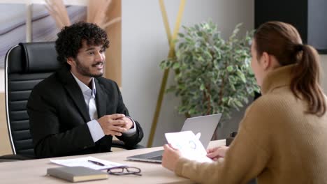 serious successful indian hr manager, entrepreneur, interviewing girl in modern office, dressed in formal suit, looking and listening to job candidate, looking at resume with concentration