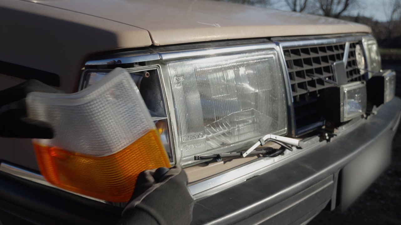 Um mecânico remove a tampa do farol dianteiro de um carro antigo para acessar a lâmpada, demonstrando um reparo prático de iluminação automotiva na parte frontal do veículo
