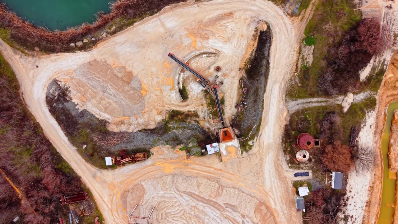 Overhead view of conveyor belt and sand stockpile at quarry site, Bulgaria. Rusty industrial equipment, white sand mound, orange-stained ground and scattered machinery.
