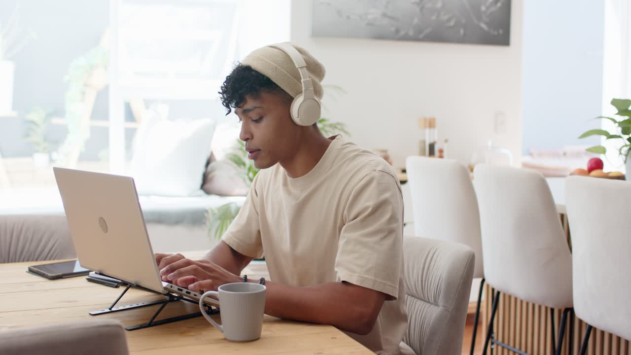 After settling at table at home, man wearing beanie, headphones typing on laptop stand for work