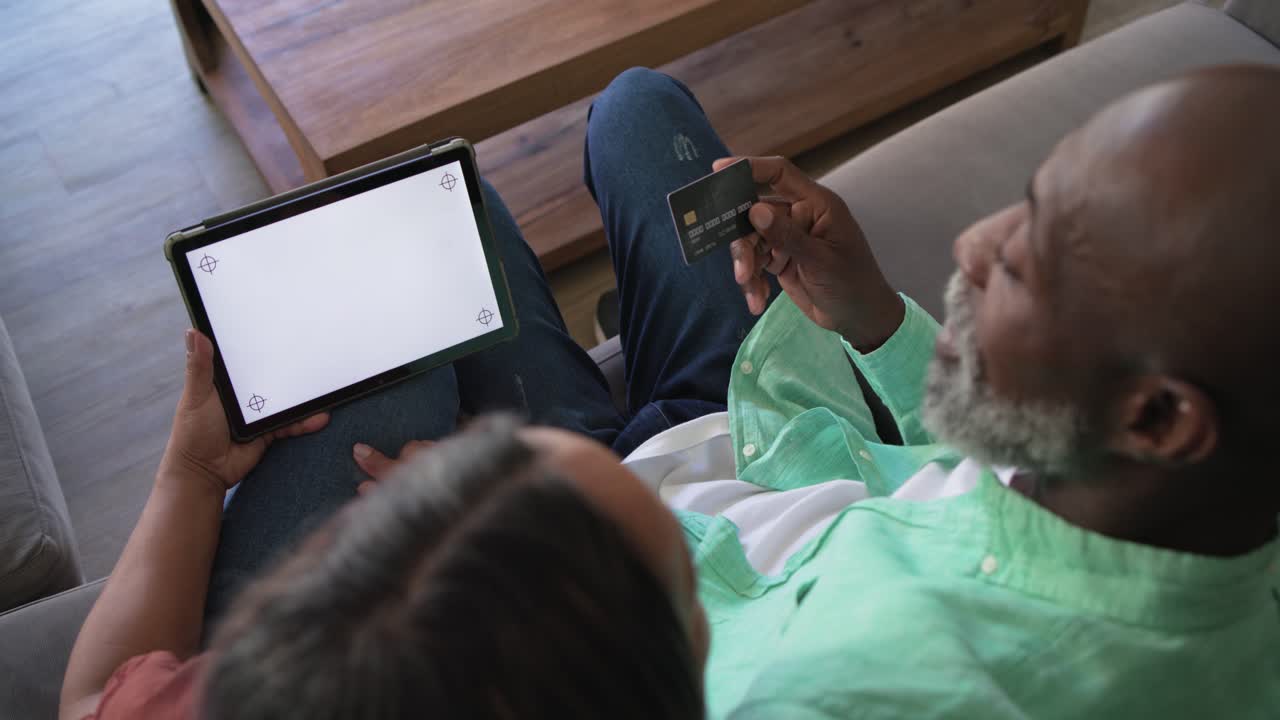 Diverse couple sitting on couch at home, blank tablet causing man angling credit card for payment