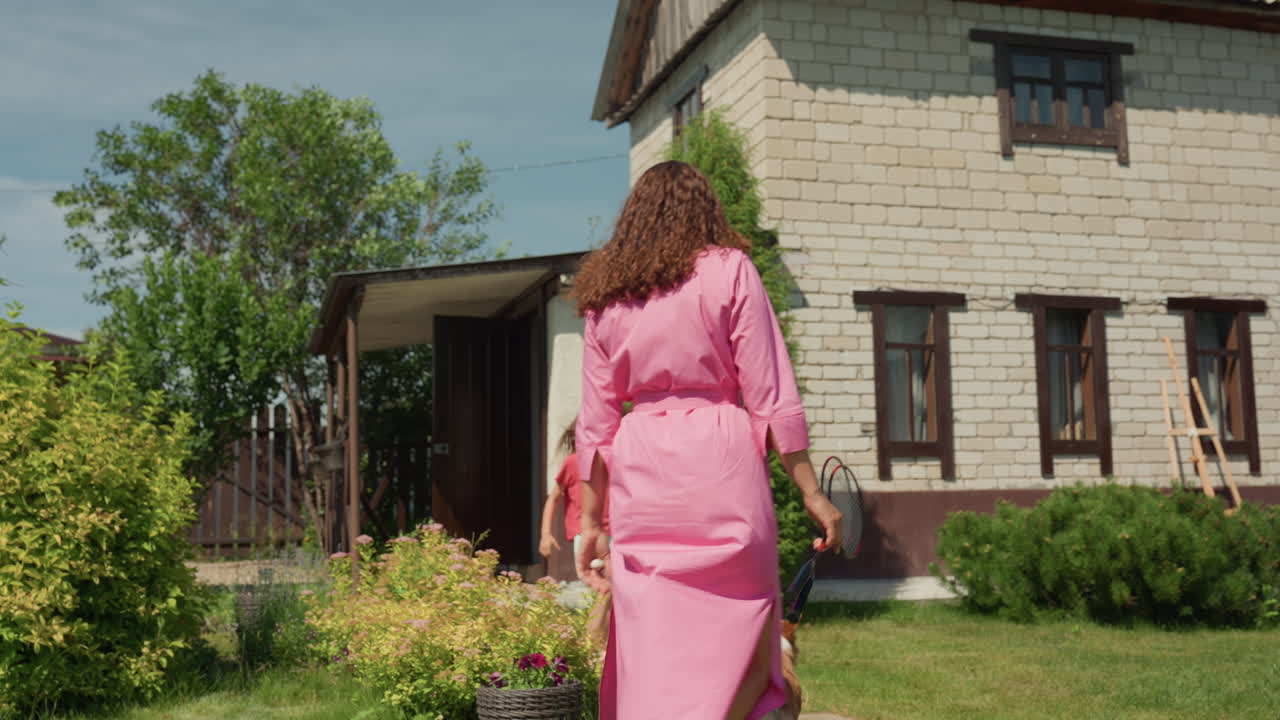Lady Approaches House With Dog And Child, Confident Woman Advances Across Yard With Playful Pet And Youngster, Cheerful Woman In Summer Attire Strolls Toward Home With Dog And Child Nearby