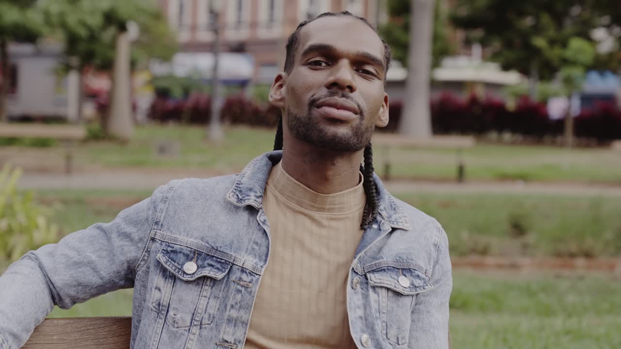 Portrait of a Black man with braided hair sitting on a bench in a park