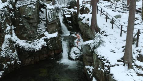 Aerial-view-of-a-person-jumping-in-a-icy-river,-at-a-waterfall,-in-middle-of-a-snowy-forest,-dark,-overcast,-winter-day---dolly,-drone-shot