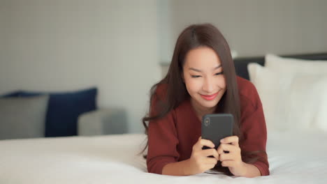 pretty asian woman using her phone with both hands typing laying on the clean bed at the hotel room wearing casual clothes close-up slow-motion