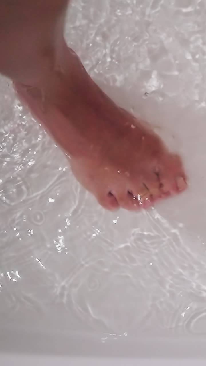 Woman foot in clear water in bathtub closeup. Bare lady leg and dripping sprays in shower cabin. Woman stands in warm liquid in home washroom