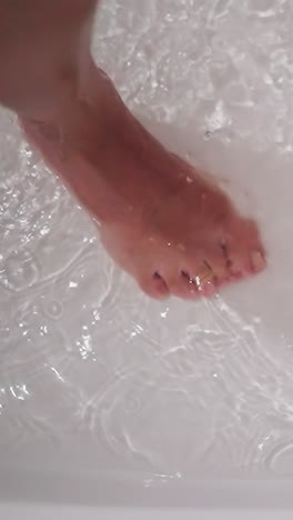 woman foot in clear water in bathtub closeup. bare lady leg and dripping sprays in shower cabin. woman stands in warm liquid in home washroom