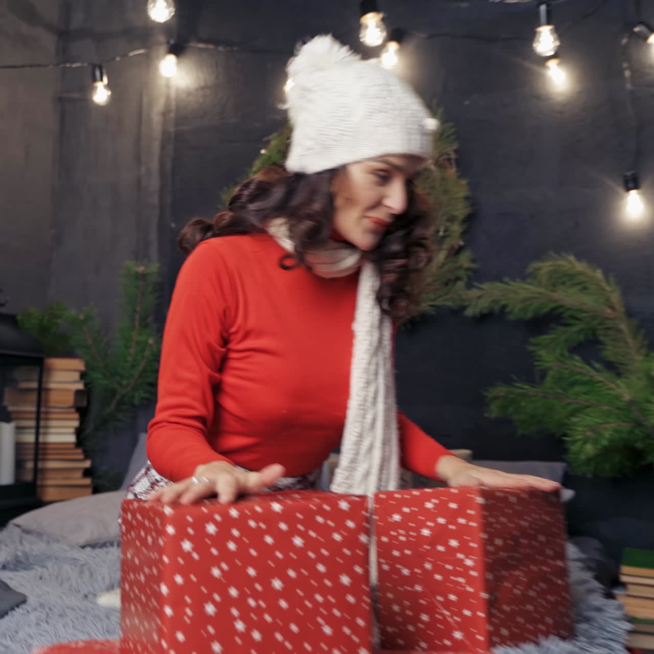 Happy woman with Christmas presents at home. Joyful girl in knitted cap and scarf sitting on bed and throwing presents on Christmas decorations background.