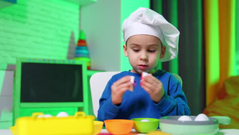 adorable toddler boy in blue sweater and white cap plays at table. cute baby chef plays with toy eggs.