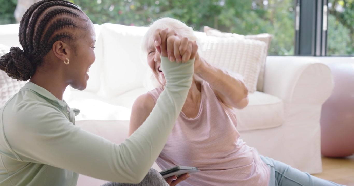 Diverse female carer and senior at home, carer showing tablet, senior tapping, smiling, learning