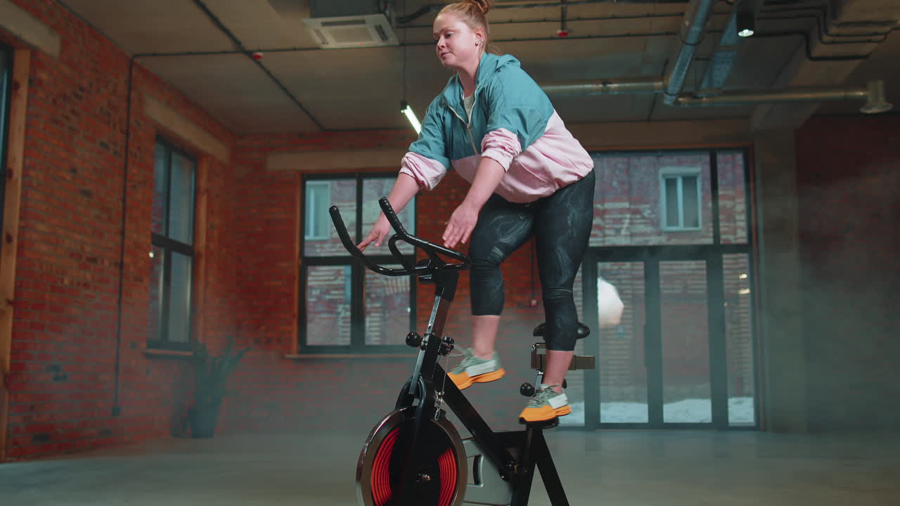 스피닝 스테이션 자전거를 타고 다니는 운동선수 여성, 안개 체육관에서 훈련 루틴, 실내에서 체중 감소