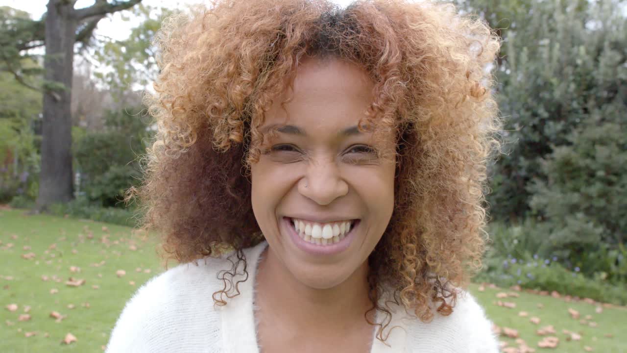 Portrait of happy african american woman wearing white cardigan, smiling in garden, slow motion