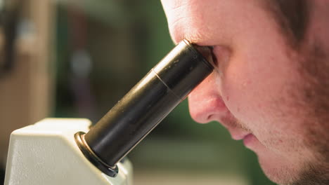 una vista de cerca de un hombre mirando atentamente a un microscopio, centrándose en su cara y el ocular, con un fondo borroso