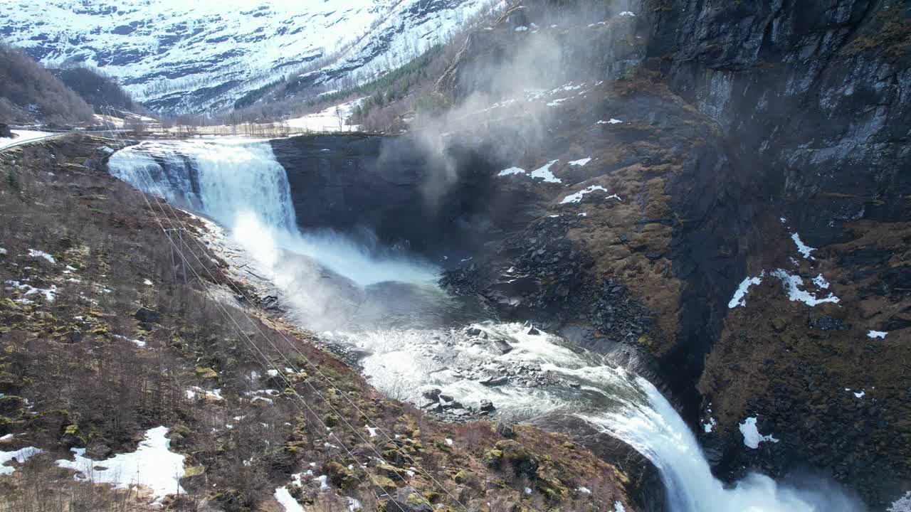 Static shot of waterfall and meltwater river in Eksingedalen. Splashing droplets, snow in background, and high voltage line show link between nature and energy