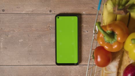 overhead studio shot of person using green screen mobile phone next to basic food items in supermarket wire shopping basket