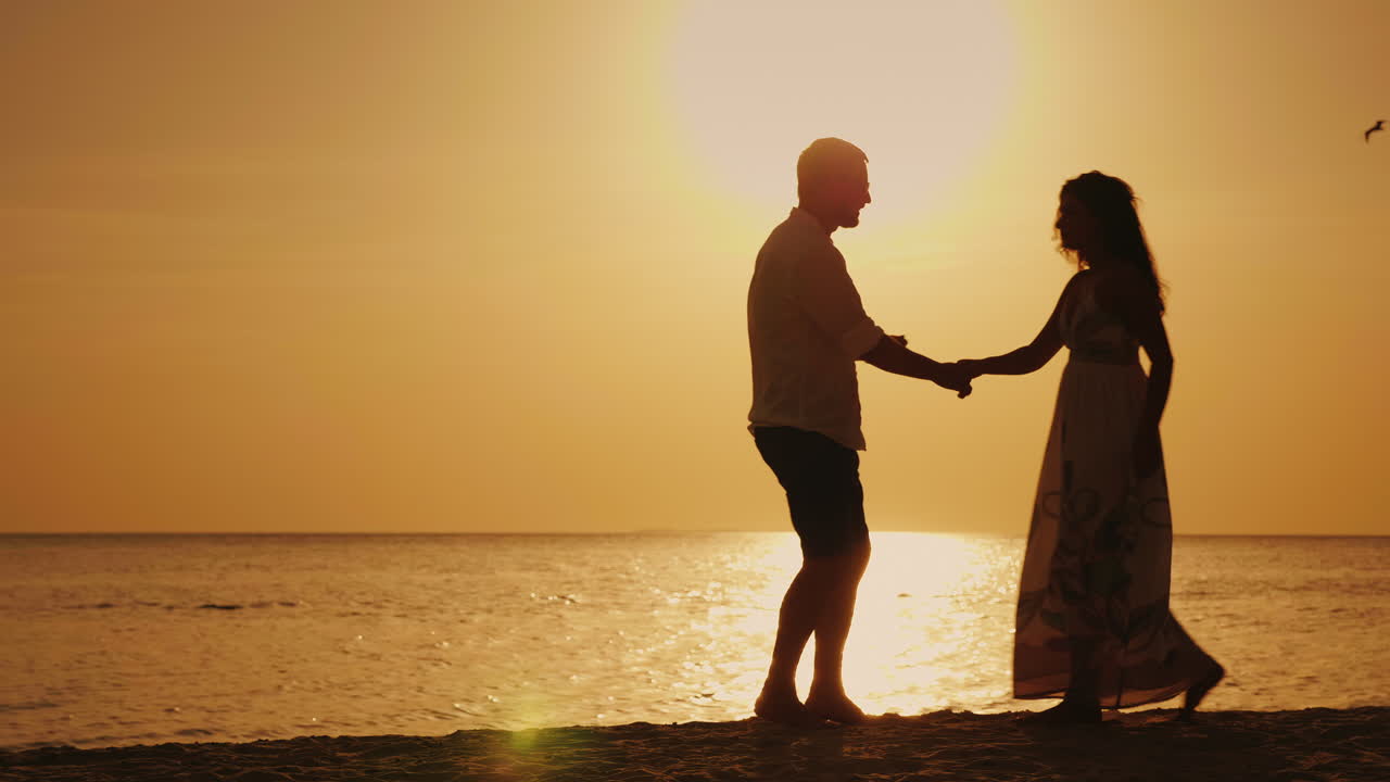 silhuetter af et ungt par, der danser mod baggrunden af havet og den glade solnedgang.
