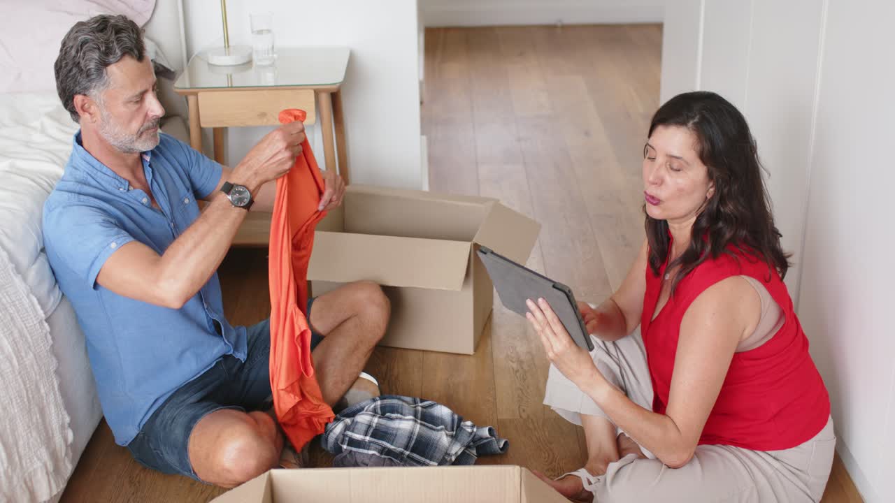 Woman consulting tablet, man is folding orange shirt and packing boxes on bedroom floor for move