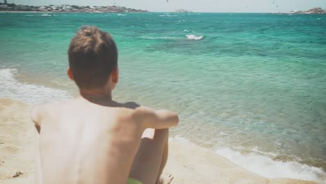 young boy sitting on a crystal beach in a greek island