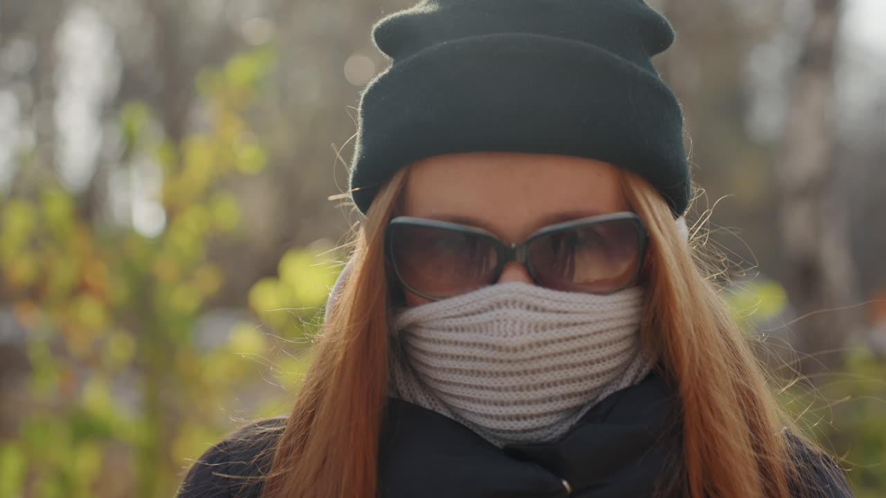 Valkoinen nainen piiloutunut huivin taakse, lähikuva silmistä ja aurinkolaseista neulotun pipon reunuksen ja vastavalon kehystämänä, pölyhiukkasia ilmassa, intensiivinen katse, kaupunkipuiston taustalla, epämiellyttävä tyyneys ja valpas jännitys