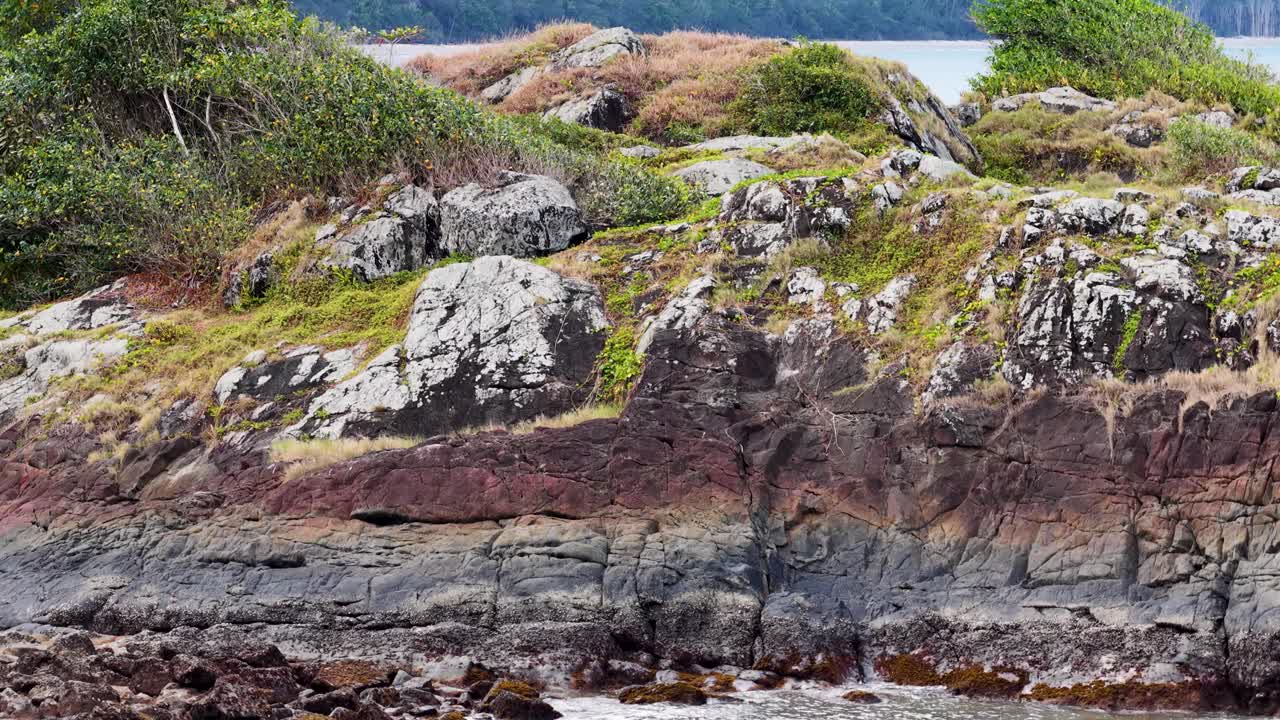 Drone camera smoothly pans across a rugged, rocky headland with green vegetation, revealing coastal cliffs and calm blue ocean under natural daylight