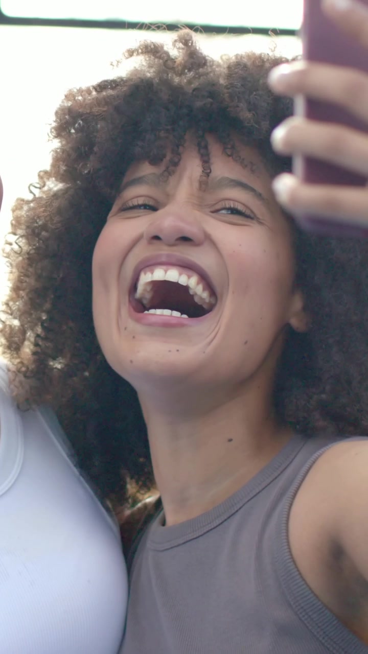Video verticale: Donna che ride, tiene in mano un telefono rosa al parco per includere un'amica che indossa un top senza maniche