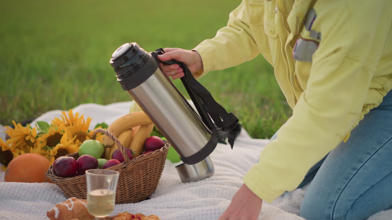 Donna inginocchiata su una coperta da picnic che apre un thermos, sistema un cesto di frutta e dei girasoli, thermos in acciaio inossidabile accanto ad anguria e croissant, che versa una bevanda calda in un bicchiere