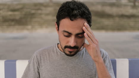 joven árabe con cabello rizado oscuro y barba en camiseta gris sentado en el muelle en un banco a rayas