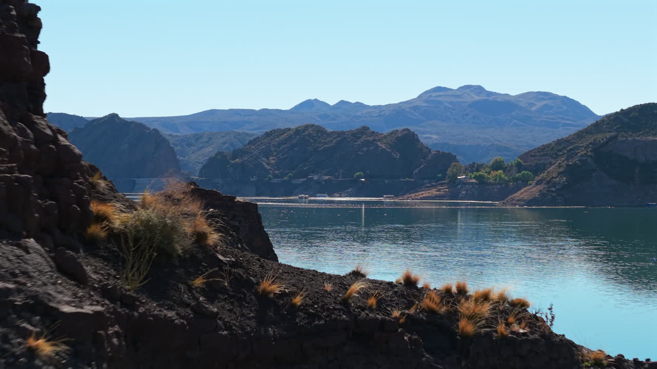 Drone reveals the water surface of the Atuel reservoir framed by dark rocky cliffs in San Rafael, Mendoza, Argentina, with the mountainous landscape in the background under bright morning light
