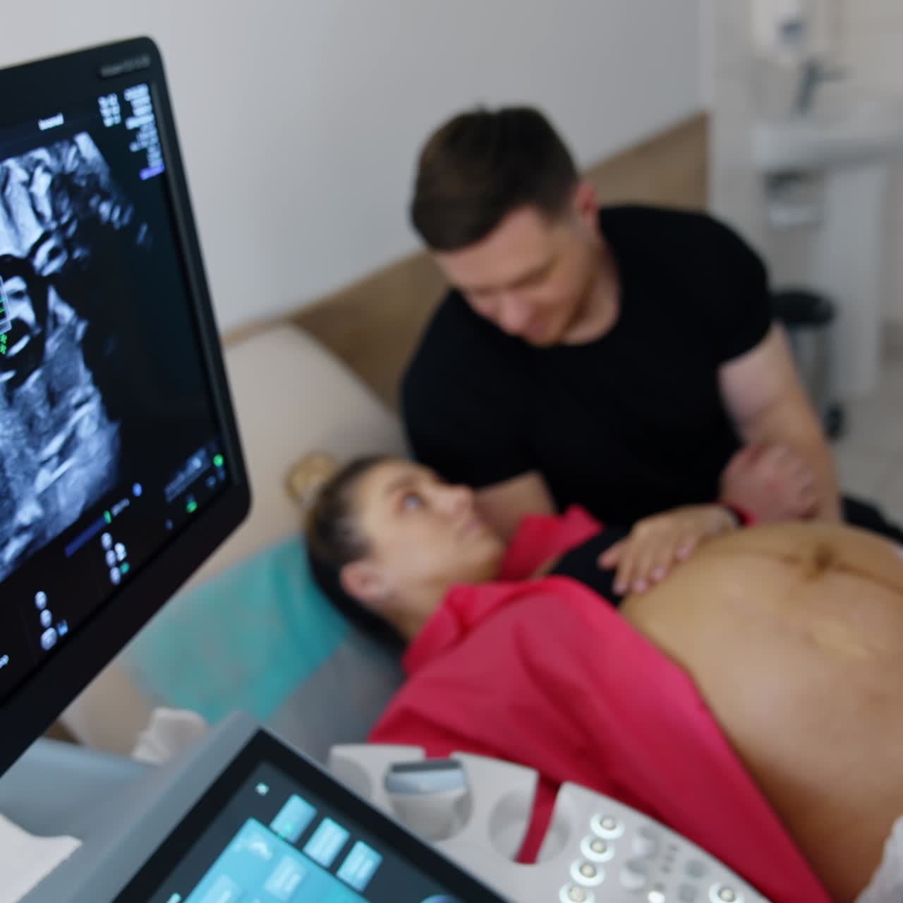 Ultrasound machine screen showing the result of pregnancy check up. Caring man kissing his expecting wife at backdrop in blur. Top view