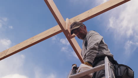 a carpenter working on a construction project stands on a ladder and then screws a small board into an overhead cross support against a bright blue sky.