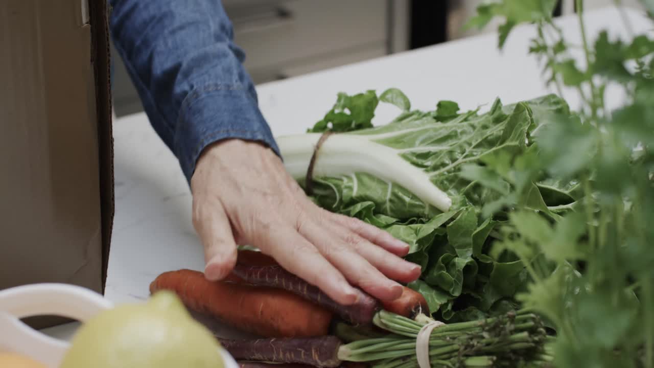 Hands of senior caucasian woman unpacking box of vegetables and fruit in kitchen, slow motion