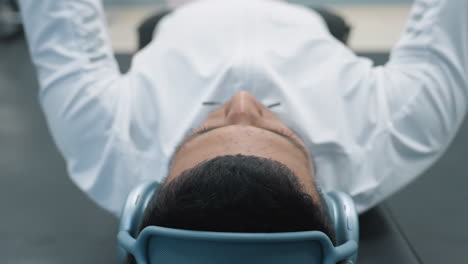 close up of man lying on bench press with headphones on, arms spread out, resting after gym workout, wearing white long sleeve shirt, soft focus background