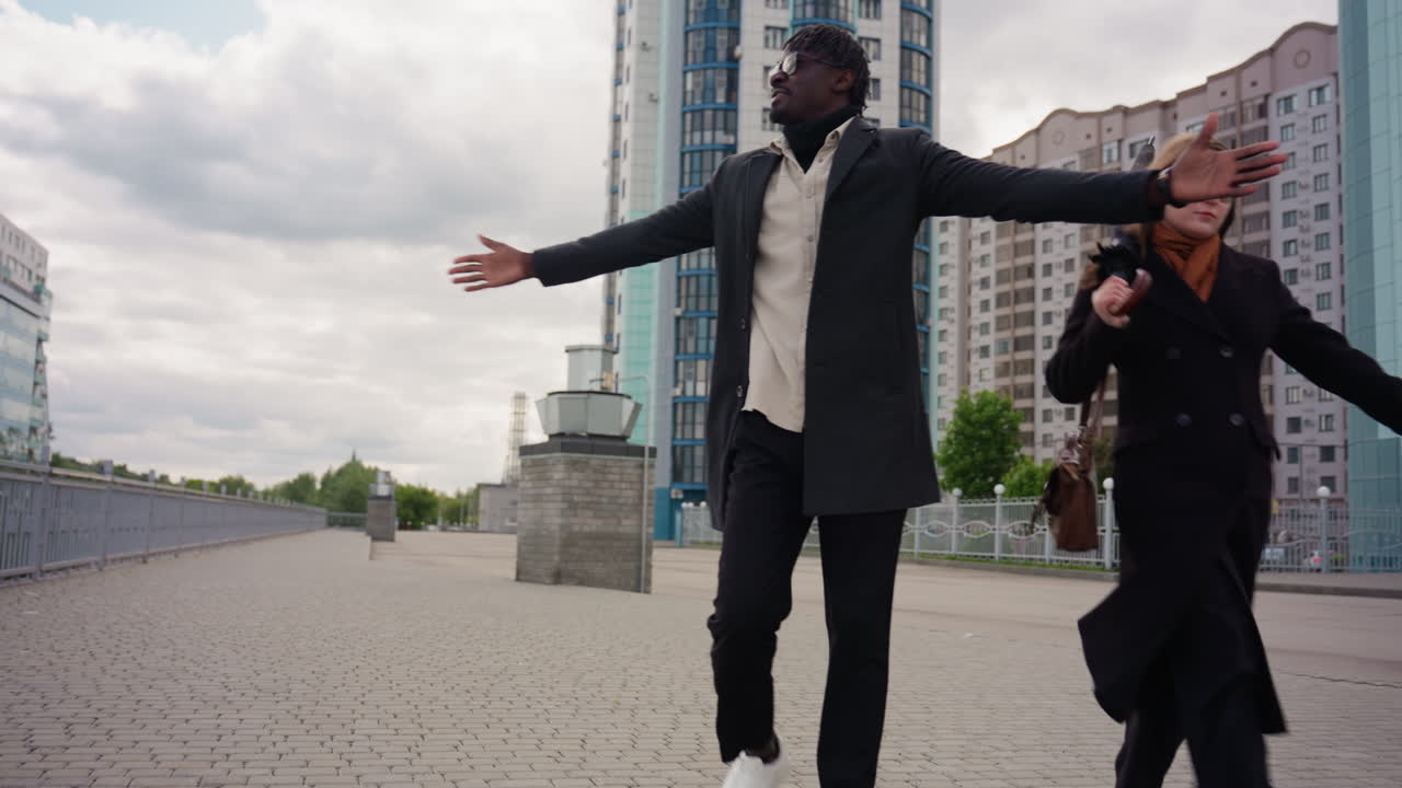 low level capturing of strolling promenade near towers, young lady flips hair backward while gentleman spreads hands wide, confident energy, urban vibe, ground level view with railing and pavement