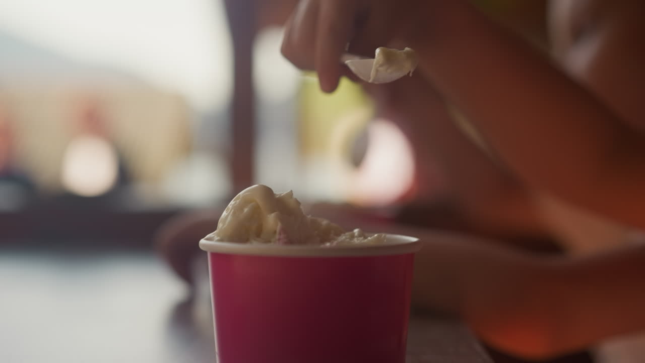 Little child hand picks vanilla ice cream with disposable spoon from paper cup sitting in cafe closeup. Kid enjoys sundae for lunch in canteen