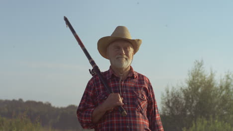 senior fisherman in a hat walking on the lake shore with a rod and equipment for fishing on a summer day