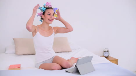 joven modelo en rodillos de cabello usando su tableta digital para conversar por video en su cama