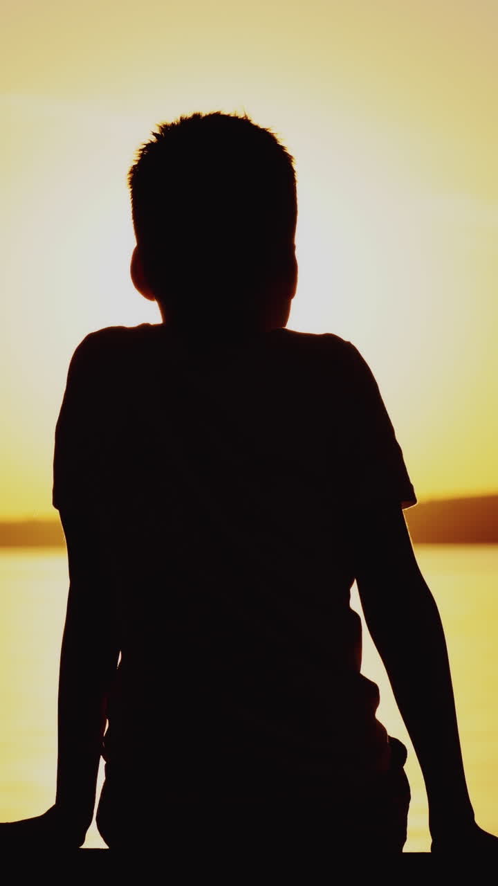 Silhouette of a boy sitting on bench at beautiful sunset over the river. Child is watching the sunset and simulates flying near the lake Vertical video